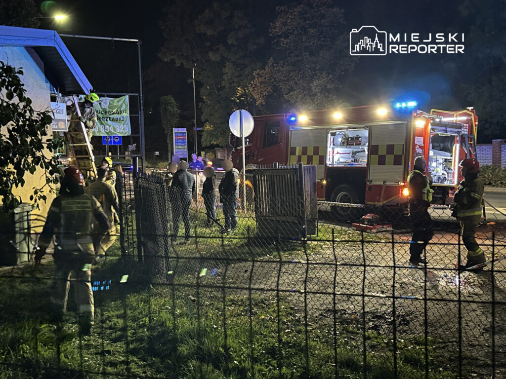 Strażacy w hełmach prowadzą działania ratunkowe przy budynku, obok wozu strażackiego z włączonymi sygnałami świetlnymi.