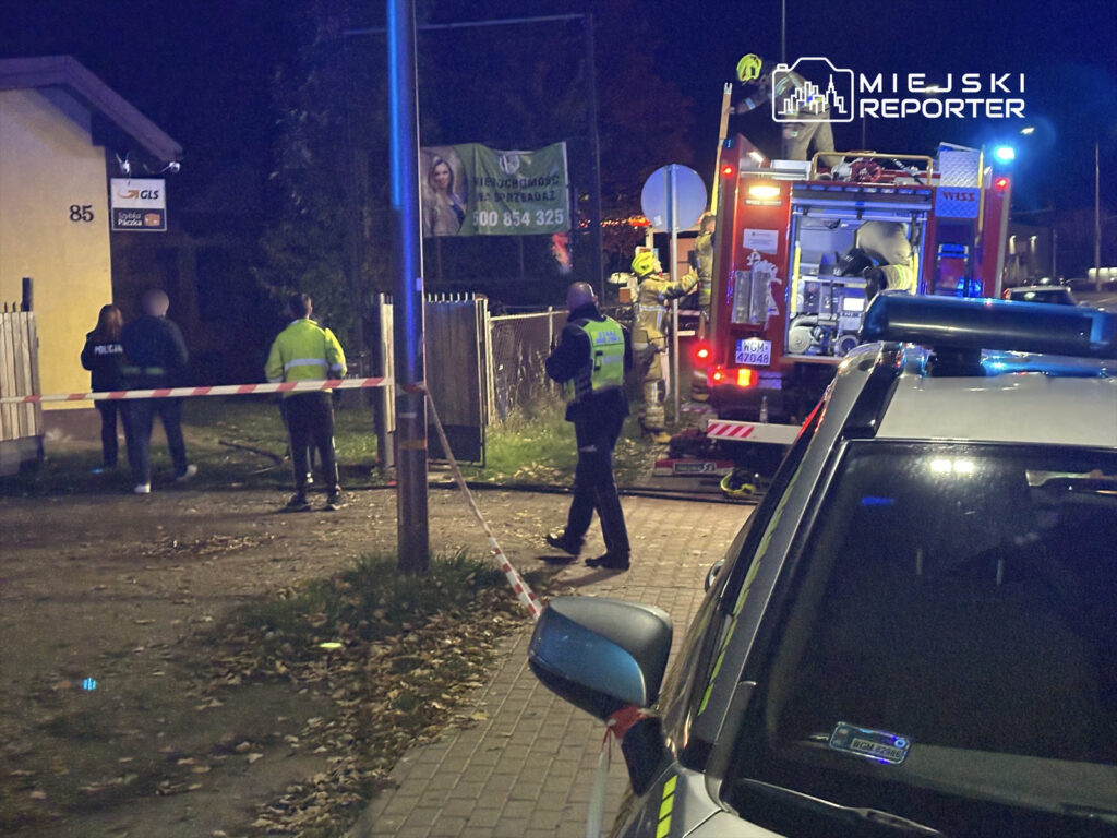 Policjanci i strażacy pracują przy zabezpieczonym terenie przed budynkiem mieszkalnym w nocy.