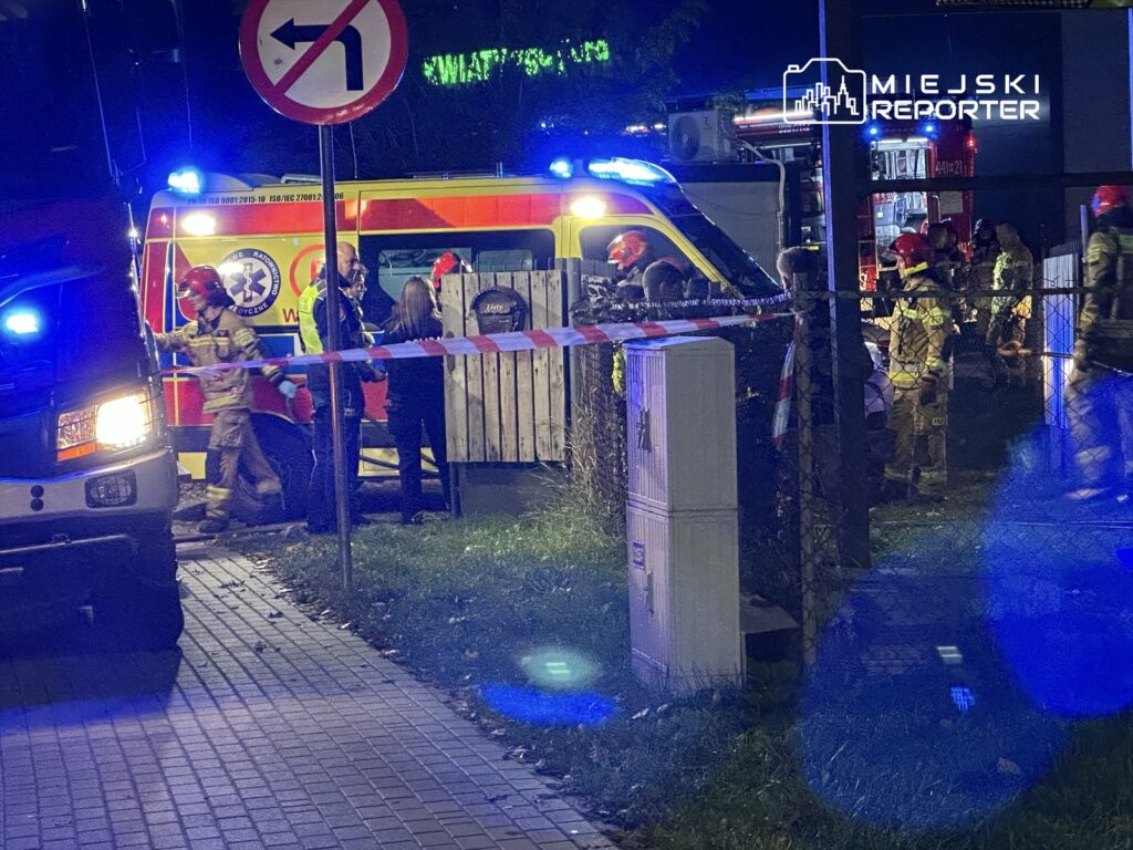 Ratownicy medyczni i strażacy pracują przy karetce w nocy, otoczeni przez barierki zabezpieczające.