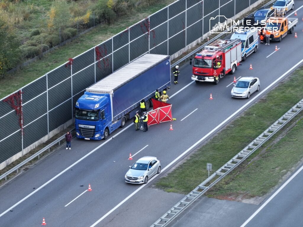Na drodze stoi niebieska ciężarówka, otoczona przez strażaków i policjantów, z włączonymi sygnałami świetlnymi.