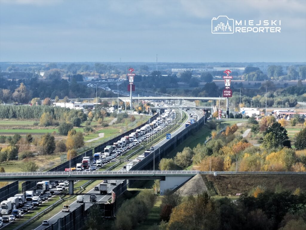 Długi korek samochodowy na autostradzie, z widocznymi ciężarówkami i stacjami benzynowymi w tle.