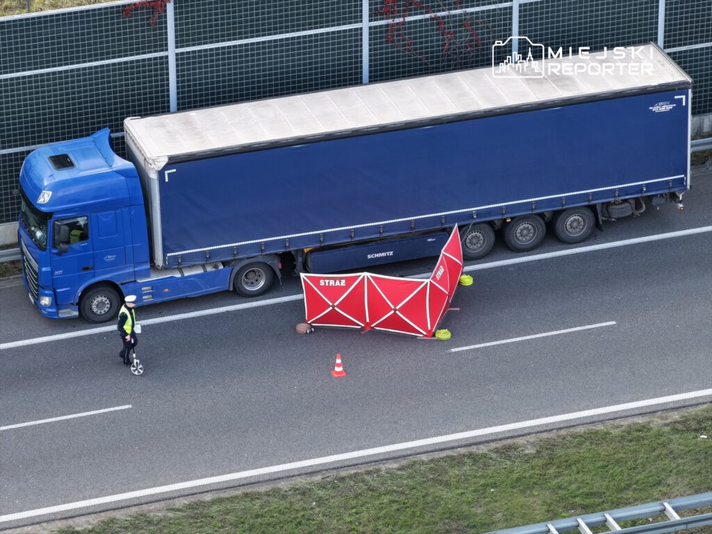 Policjant w kamizelce odblaskowej mierzy odległość obok niebieskiego tira, a w tle znajduje się czerwony namiot strażacki.