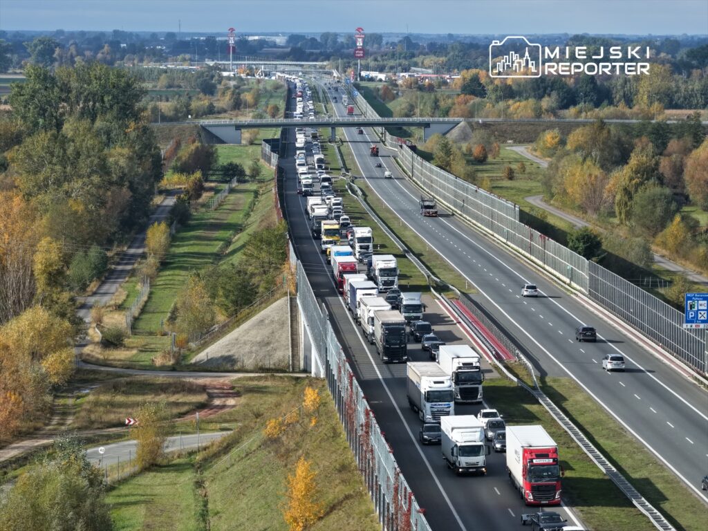 Kolejka samochodów ciężarowych i osobowych na zatłoczonej autostradzie, w otoczeniu drzew i zieleni.