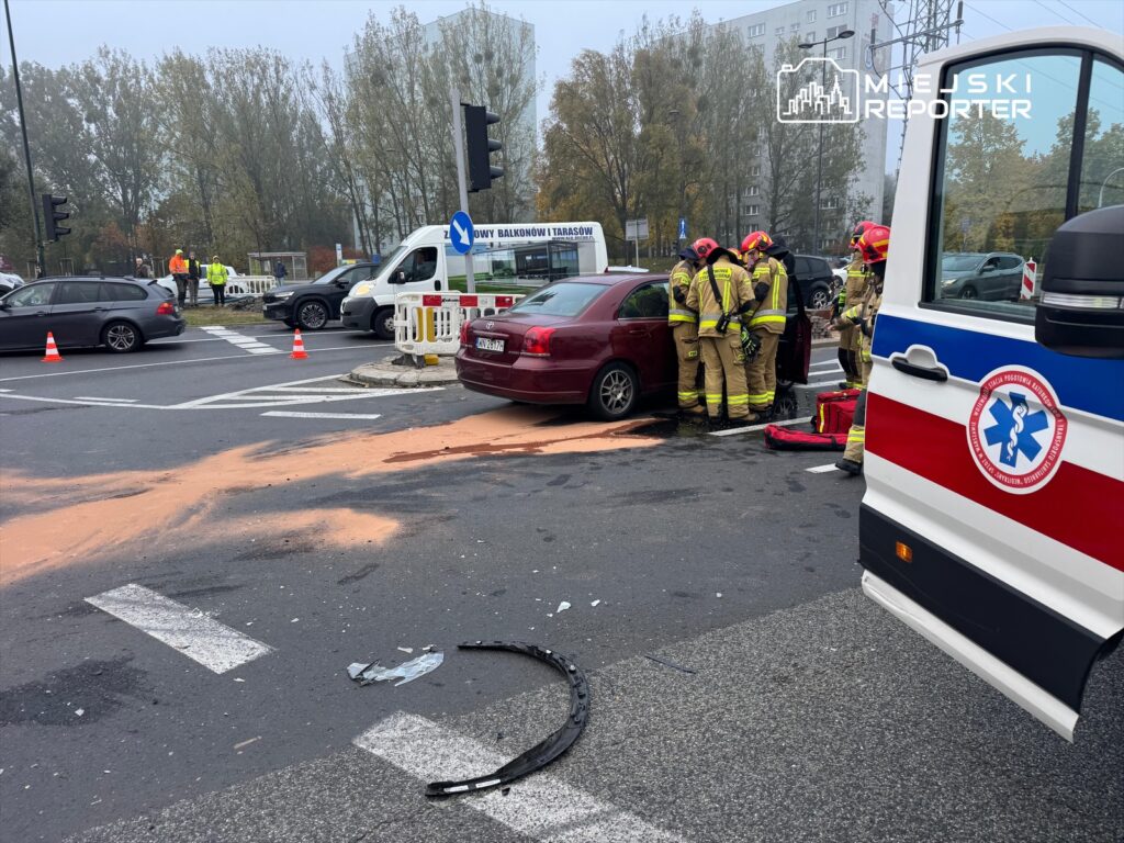 Grupa strażaków udziela pomocy poszkodowanemu w czołowej kolizji na skrzyżowaniu, w tle widać inne pojazdy.