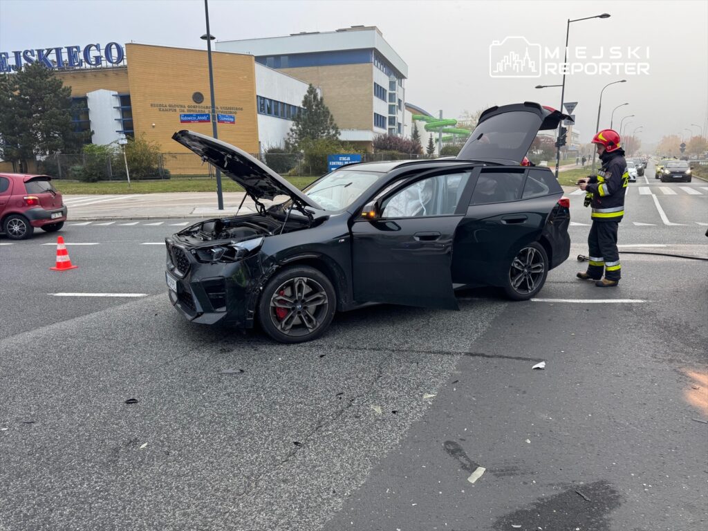 Czarny samochód osobowy z uszkodzoną przednią częścią stoi na drodze, obok strażak w mundurze sprawdza sytuację.