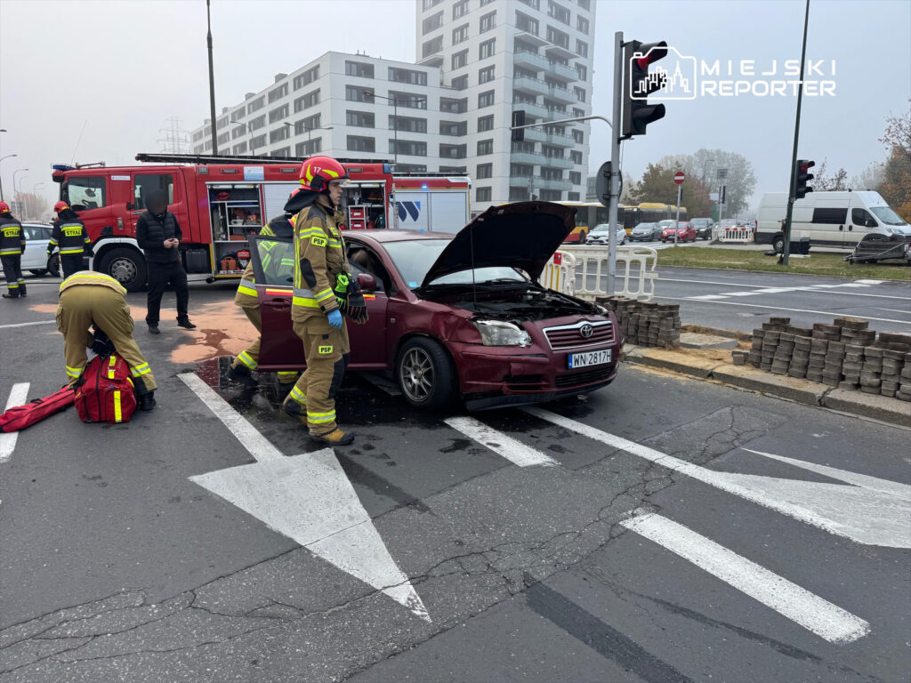 Strażacy w mundurach interweniują przy uszkodzonym samochodzie osobowym z otwartą maską, na ruchliwej ulicy.
