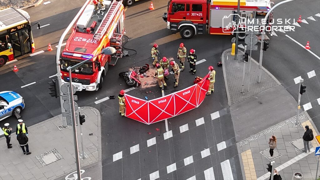 Grupa strażaków zabezpiecza miejsce wypadku drogowego, wokół leży przewrócony motocykl, a w tle widać pojazdy służb ratunkowych.