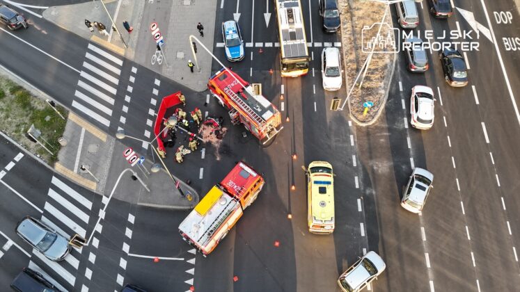 Zespół ratunkowy udziela pomocy poszkodowanym na skrzyżowaniu, obok dwóch wozów strażackich i karetki.