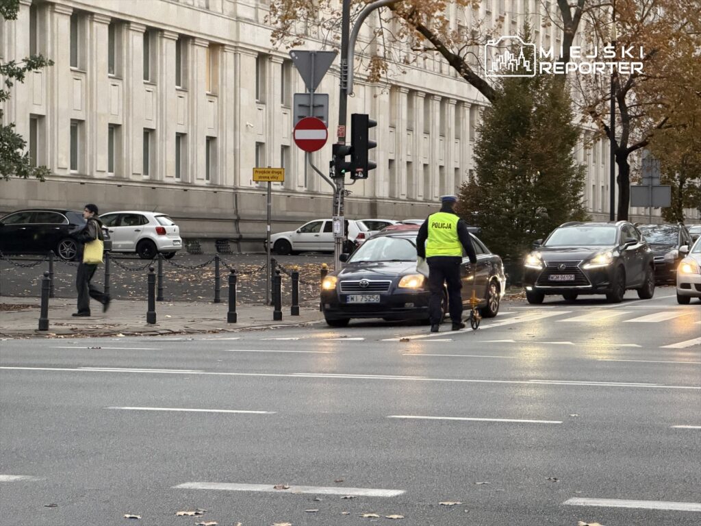 Policjant w kamizelce odblaskowej mierzy prędkość pojazdów na ulicy, obok przechodnia z torbą.