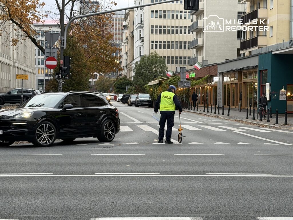 Policjant w kamizelce odblaskowej mierzy odległość na jezdni w miejskim otoczeniu, obok zaparkowanych samochodów.