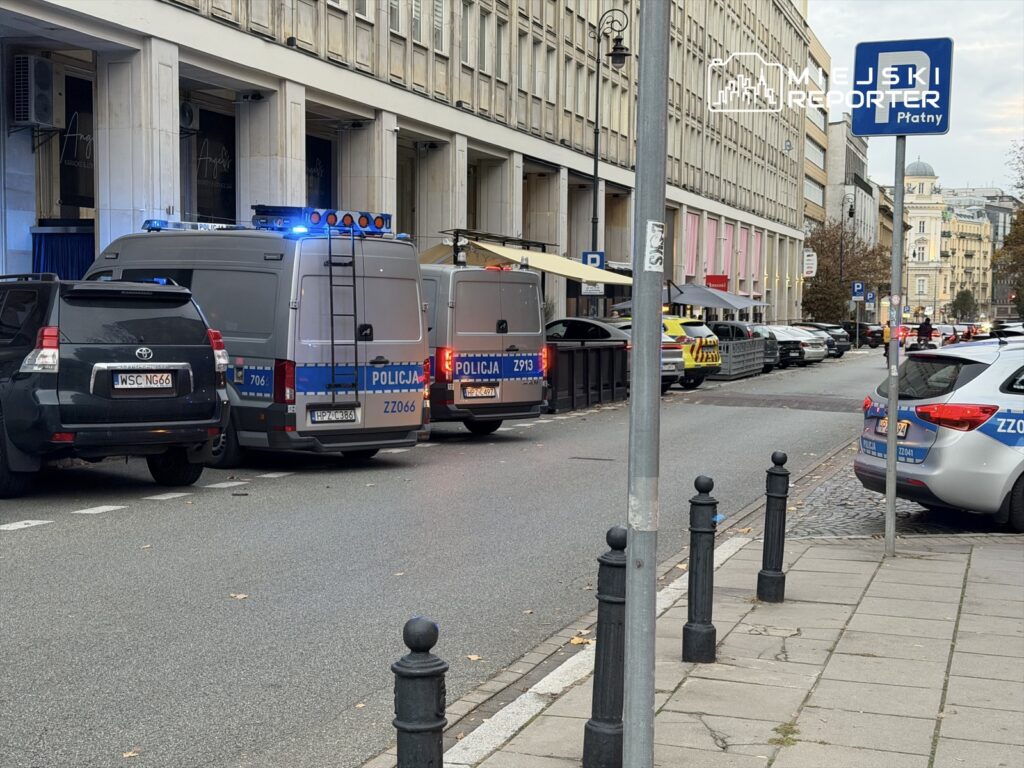 Na ulicy parkują pojazdy policyjne z włączonymi sygnałami świetlnymi, obok stojący czarny SUV.