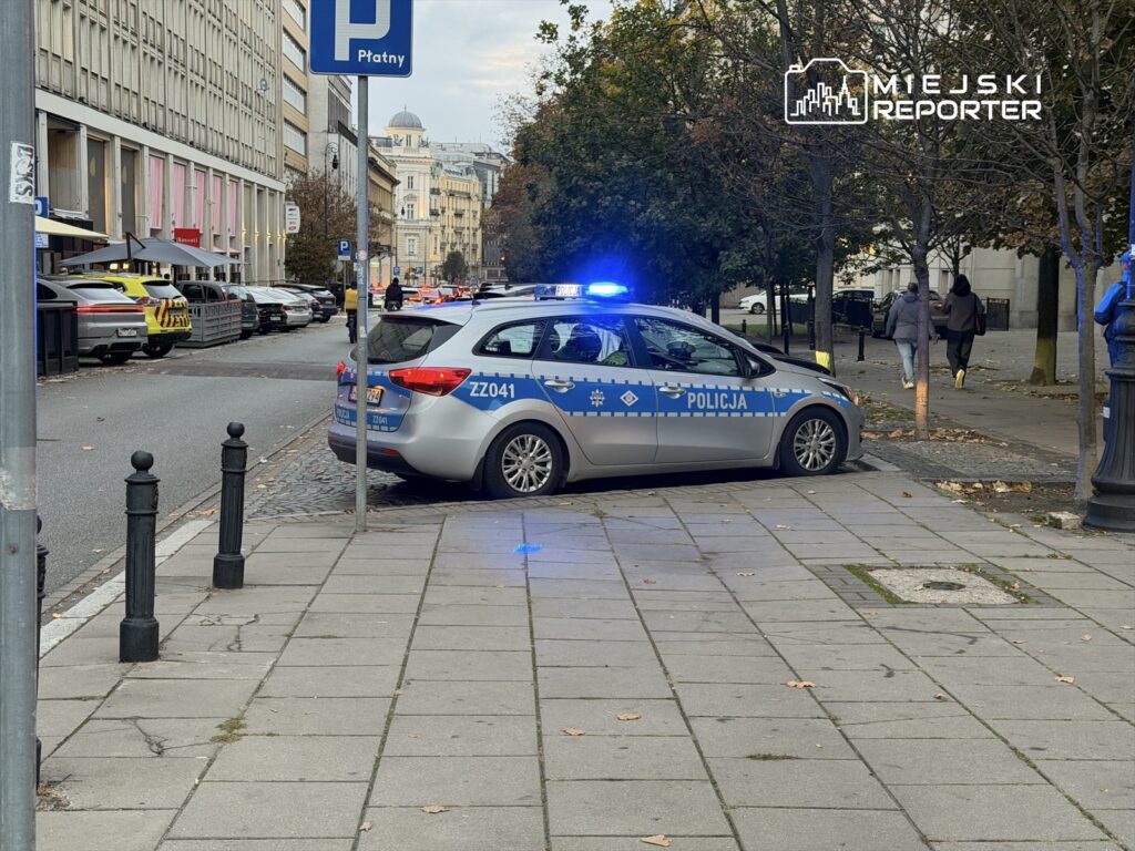 Policyjny radiowóz zaparkowany na chodniku, z włączonymi sygnałami świetlnymi, obok ulicy z samochodami.