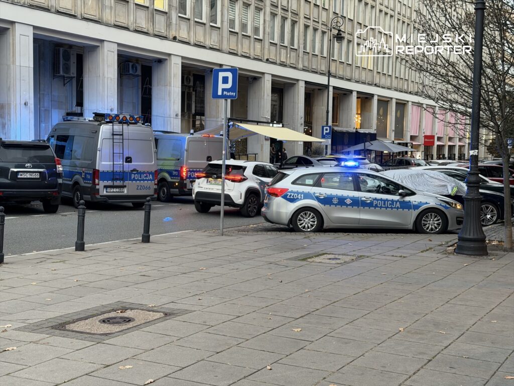 Policyjny radiowóz z włączonymi sygnałami świetlnymi stoi na ulicy, obok kilku innych samochodów i zaparkowanych furgonetek.