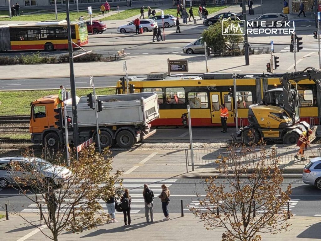 Pomarańczowy ciężarówka z wywrotką stoi obok żółtego tramwaju, a pracownicy w kamizelkach odblaskowych prowadzą prace na torach.