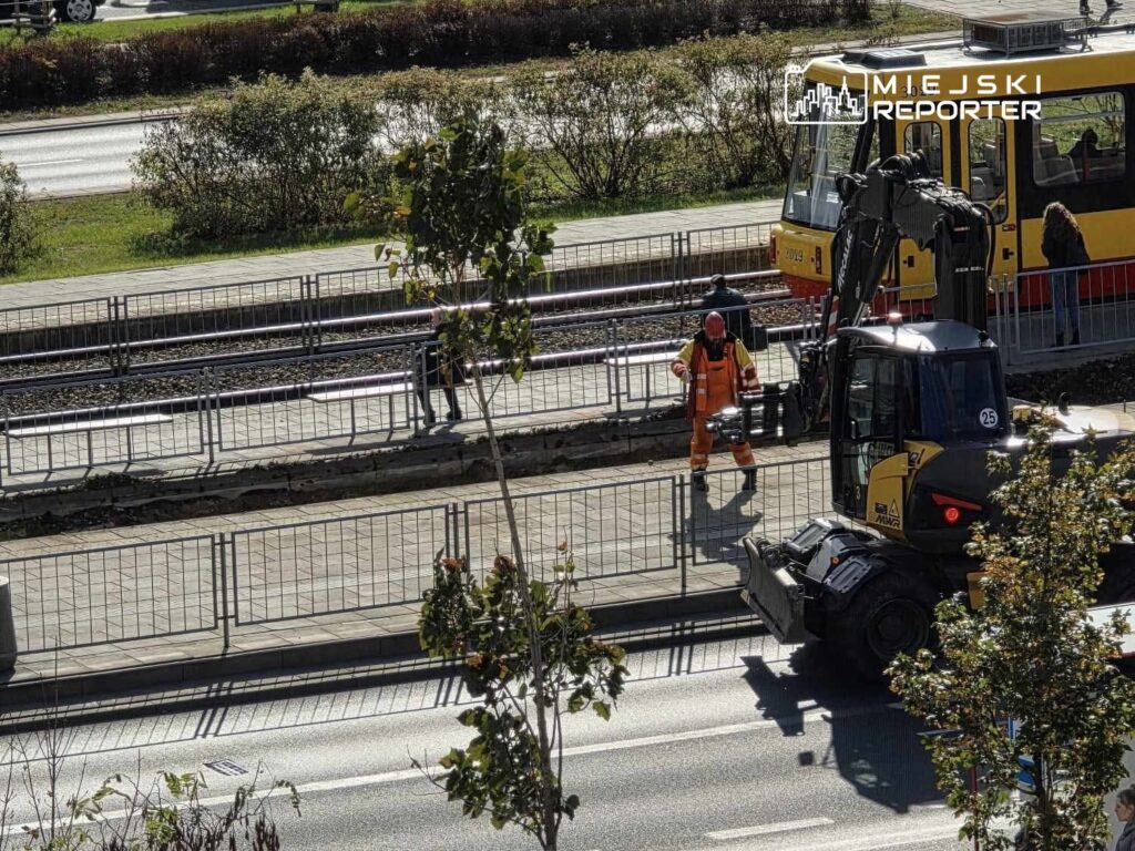Robotnik w pomarańczowym uniformie pracuje przy torach tramwajowych, obok przystanku z tramwajem.