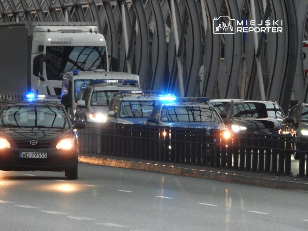 Policyjny radiowóz z niebieskimi syrenami porusza się w zatorze drogowym, obok ciężarówki i innych samochodów.