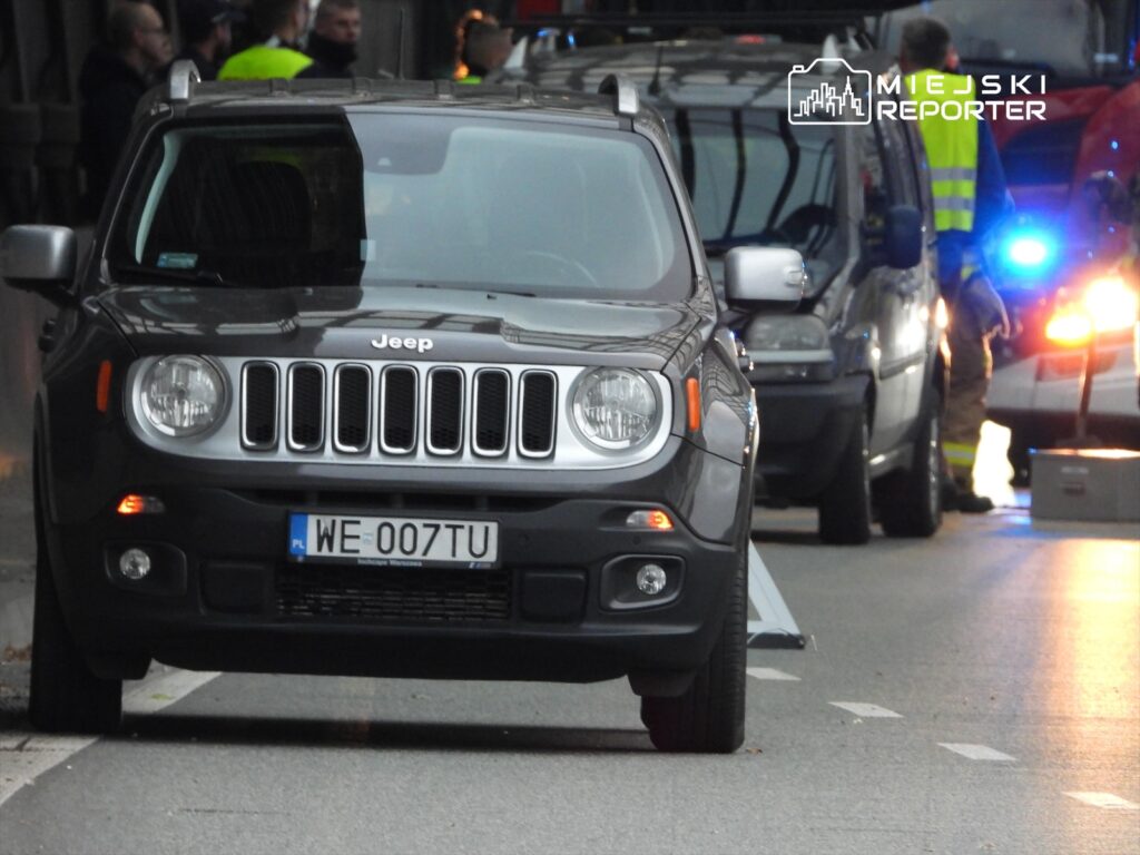 Czarny SUV marki Jeep zaparkowany na ulicy, w tle widać funkcjonariuszy oraz pojazdy służb ratunkowych.