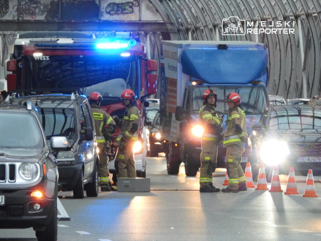 Strażacy w hełmach omawiają sytuację na zablokowanej drodze, obok zaparkowanej ciężarówki i samochodów osobowych.