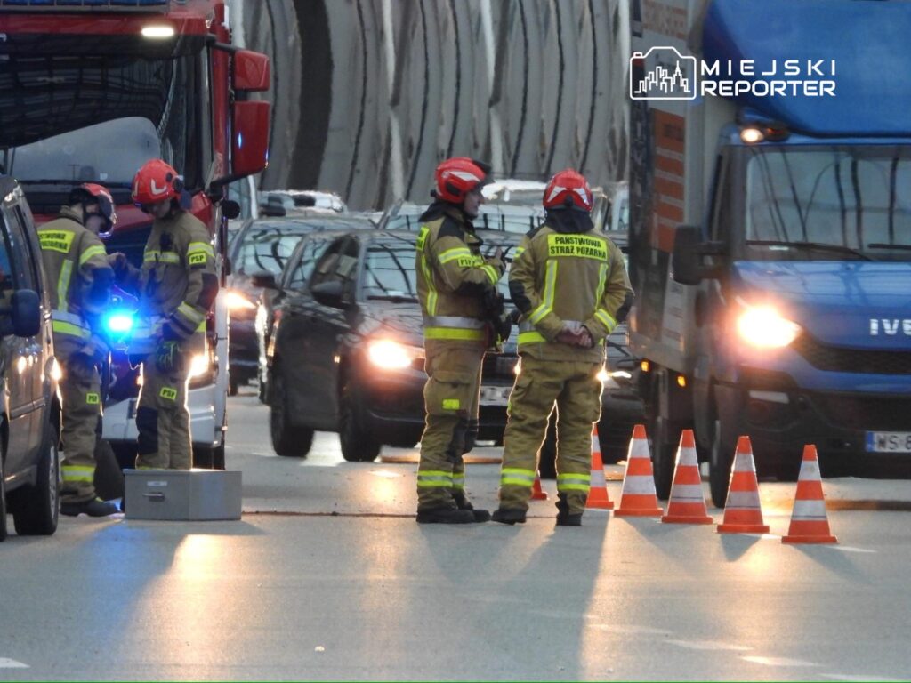 Grupa strażaków w mundurach omawia sytuację na zablokowanej drodze, obok wozu strażackiego oraz ustawionych pachołków.