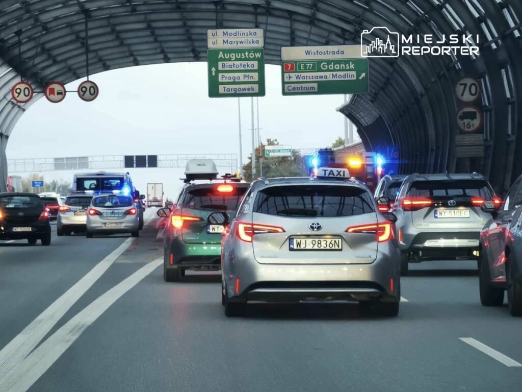 Kolejka samochodów zatrzymana na drodze, w tle widoczne znaki kierunkowe i pojazdy z włączonymi sygnalizacjami świetlnymi.