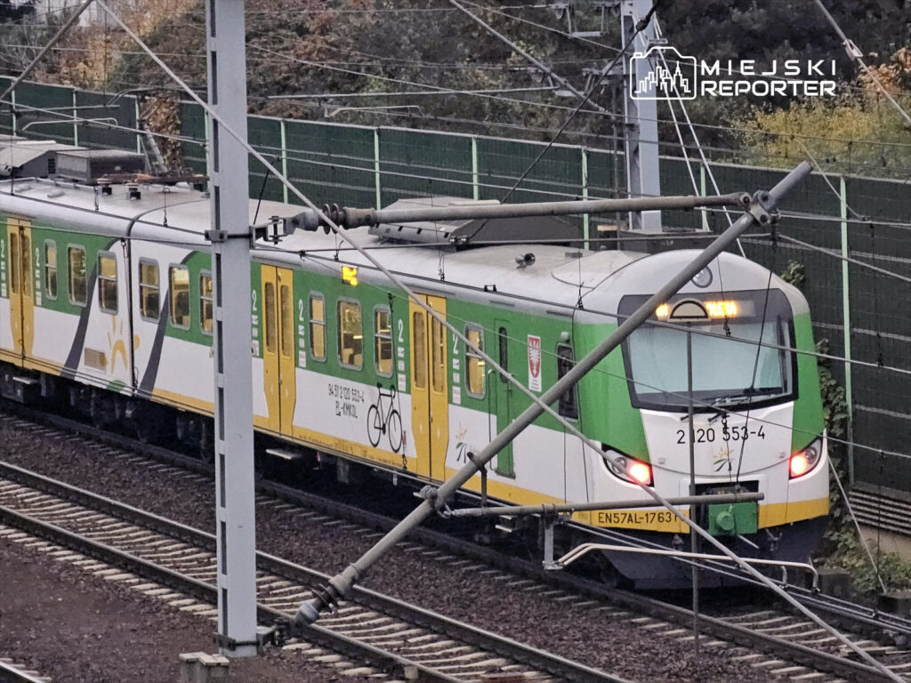 Pociąg elektryczny w zielono-żółtych barwach zatrzymuje się na stacji w otoczeniu torów i ogrodzenia.