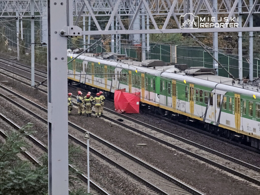 Grupa strażaków w hełmach zbiera się przy torach kolejowych obok pociągu, zasłoniętego czerwoną plandeką.
