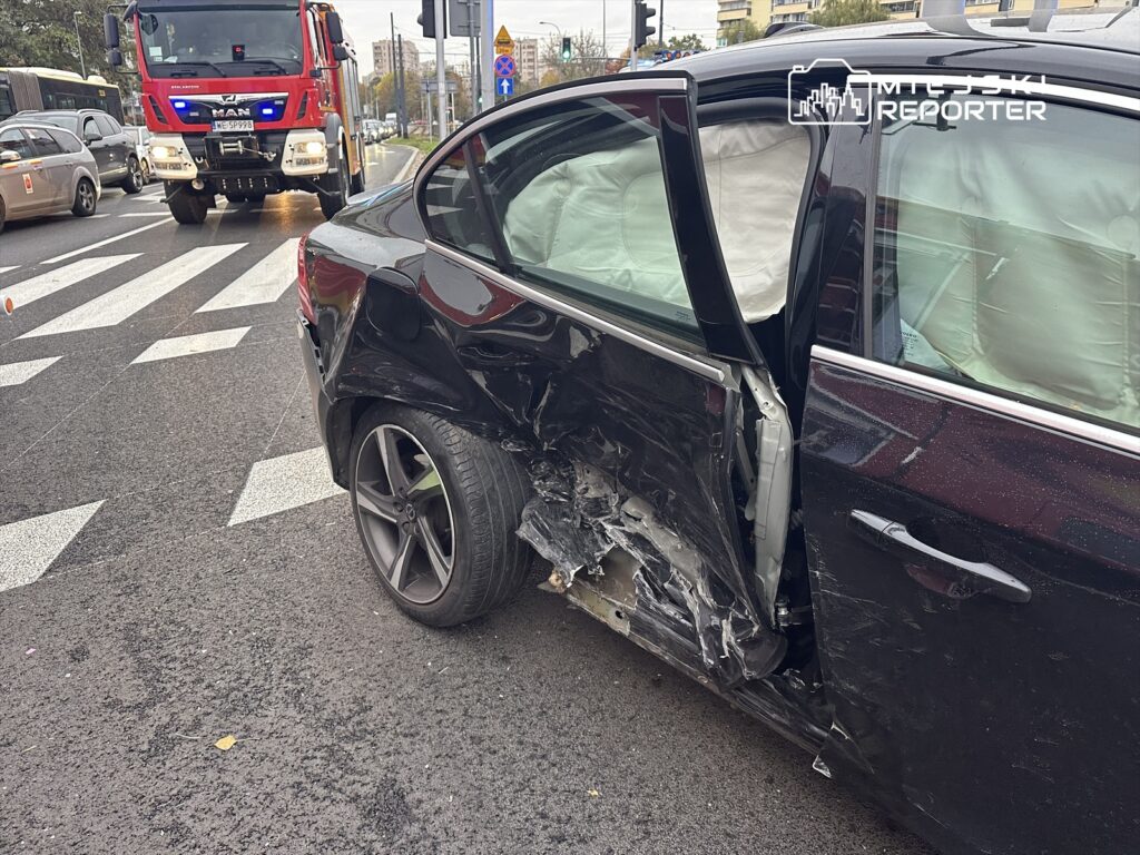 Czarny samochód osobowy z uszkodzoną lewą stroną stoi na jezdni, obok przejeżdża wóz strażacki.