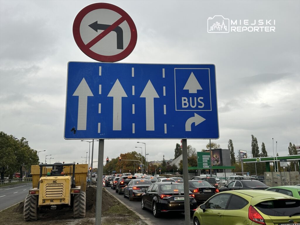 Znak drogowy zakazujący skrętu w lewo oraz wskazujący kierunek jazdy dla pojazdów i autobusów. W tle widoczny korek samochodowy.