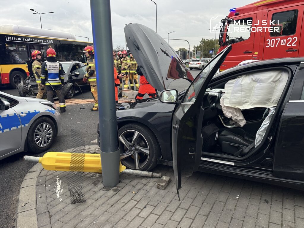 W miejscu wypadku drogowego strażacy i ratownicy medyczni udzielają pomocy poszkodowanym, przy uszkodzonym samochodzie.