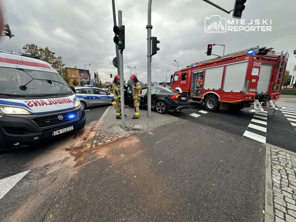 Karetka pogotowia oraz wóz strażacki zatrzymane przy skrzyżowaniu, a strażacy rozmawiają obok uszkodzonego samochodu.