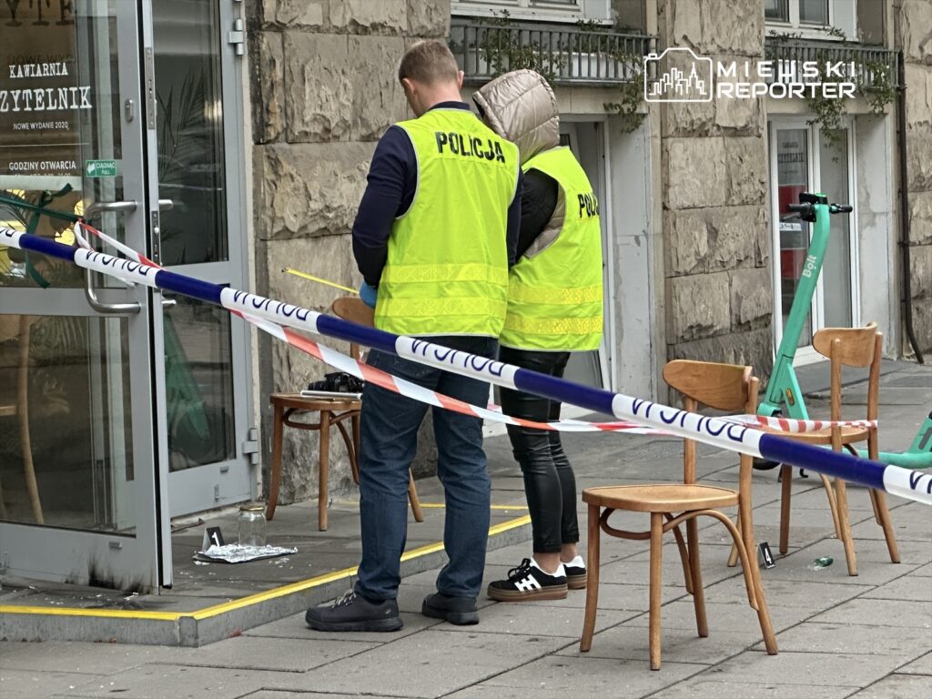 Policjanci w kamizelkach odblaskowych prowadzą czynności na miejscu zdarzenia przed kawiarnią, obok pozostawione krzesła.