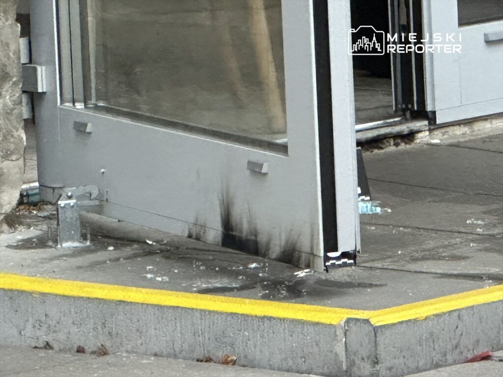 Zniszczona szklana drzwi z widocznymi śladami po pożarze, stojąca na chodniku w miejskim otoczeniu.