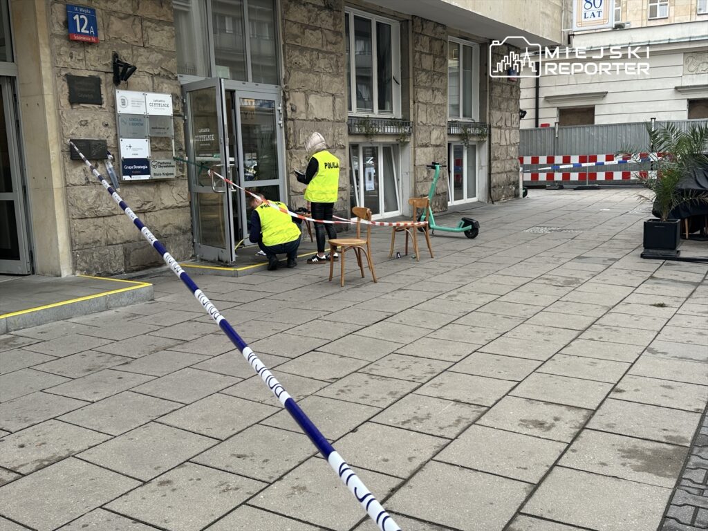 Funkcjonariusze policji zabezpieczają wejście do budynku, obok znajdują się krzesła i hulajnoga.