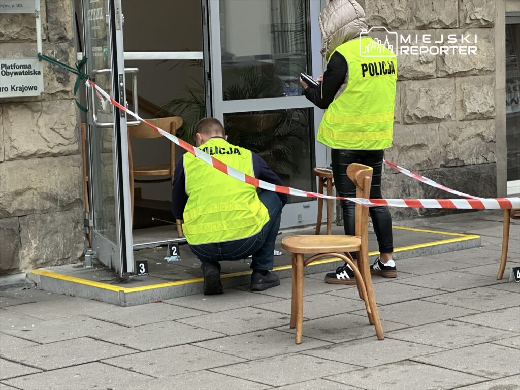 Dwóch policjantów w kamizelkach odblaskowych bada miejsce zdarzenia przed wejściem do budynku, zabezpieczonym taśmą.