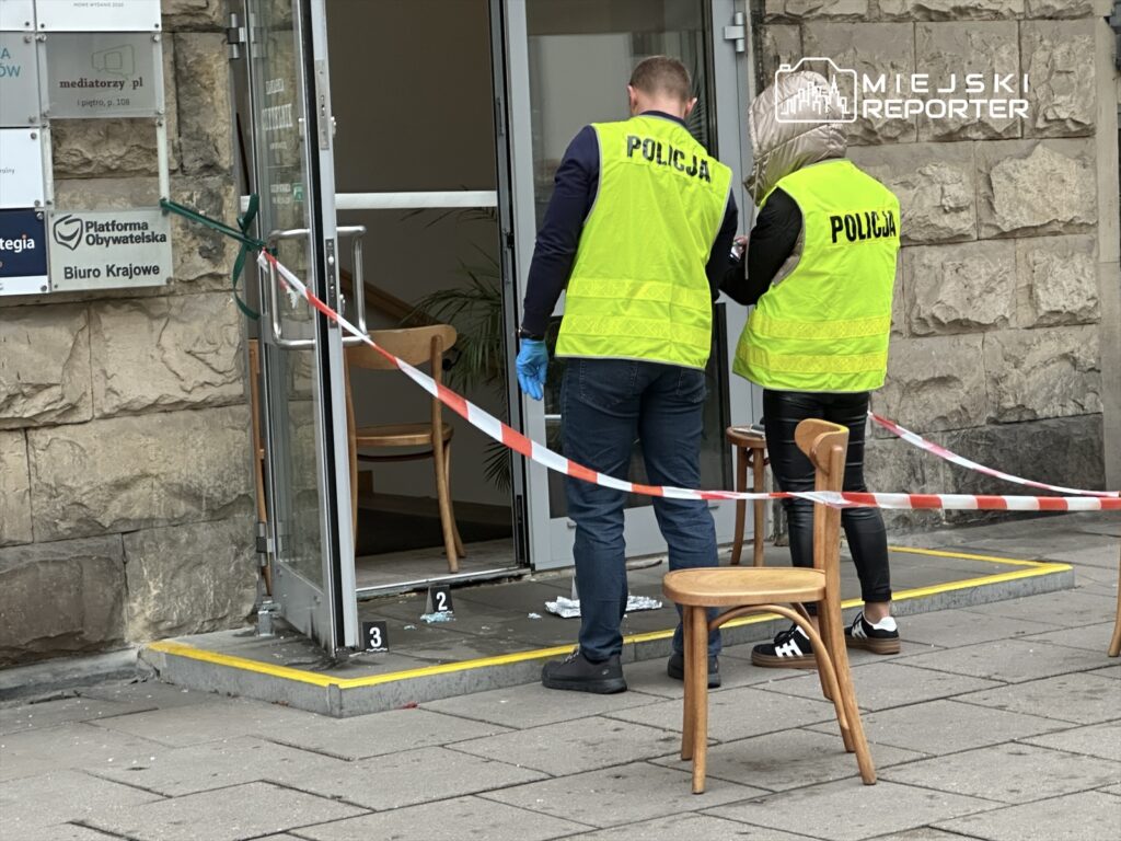 Policjanci w kamizelkach odblaskowych badają miejsce zdarzenia przed wejściem do biura, zabezpieczając teren taśmą.