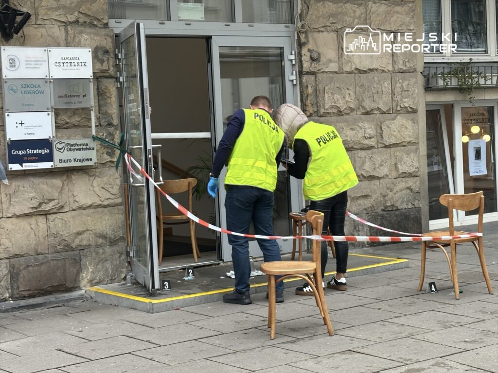 Policjanci w kamizelkach odblaskowych badają miejsce zdarzenia przed budynkiem z oznaczeniami firmowymi.
