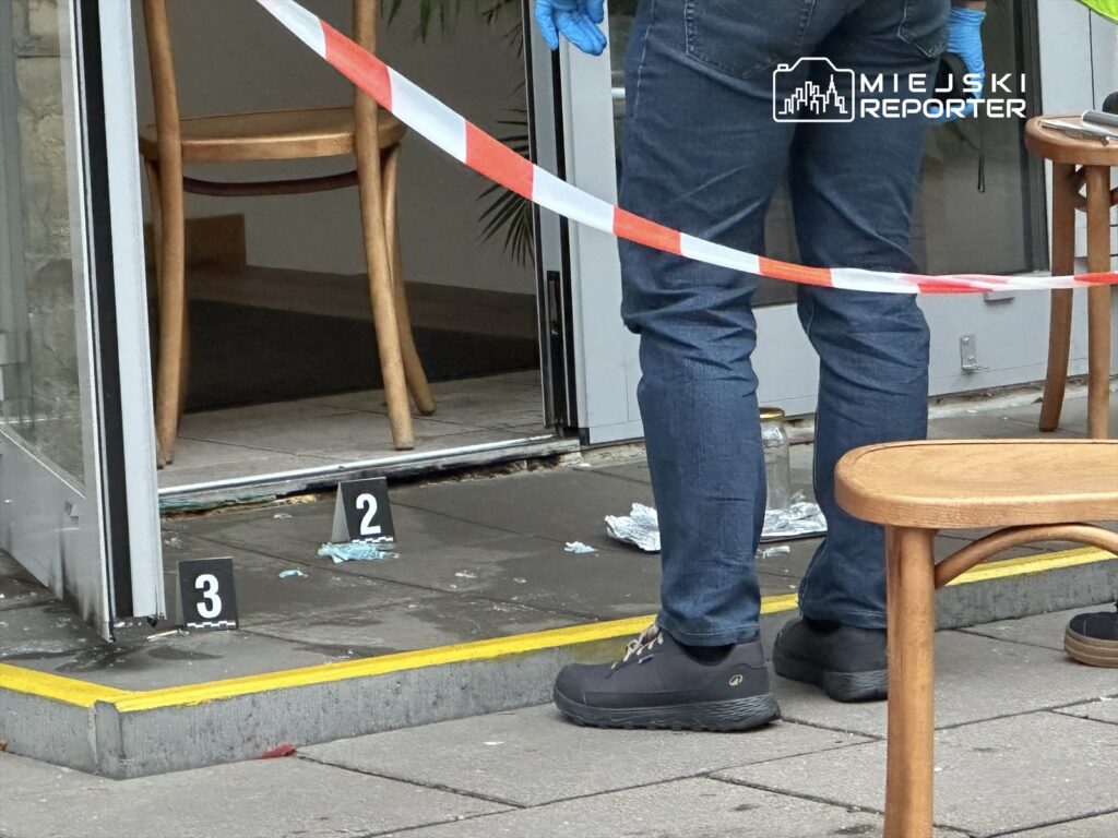 Mężczyzna w niebieskich rękawiczkach stoi przy wejściu do lokalu, zabezpieczając miejsce taśmą ostrzegawczą.