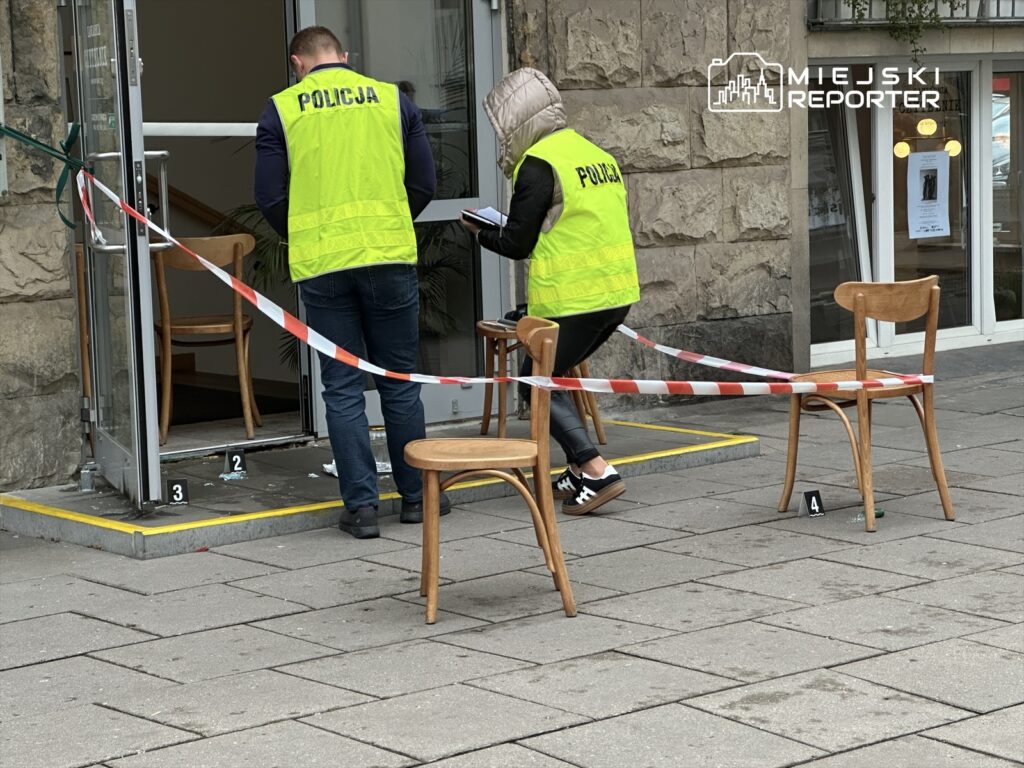 Funkcjonariusze policji w kamizelkach odblaskowych zbierają dowody przed wejściem do budynku, zabezpieczając teren taśmą.