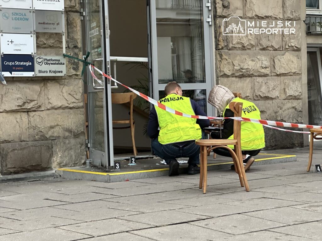 Policjanci w żółtych kamizelkach zabezpieczają miejsce zdarzenia przed wejściem do biura.