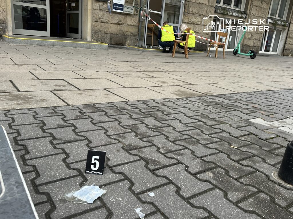 Na chodniku przed budynkiem dwie osoby w żółtych kamizelkach zabezpieczają miejsce zdarzenia, obok leży oznaczenie „5” oraz fragment szkła.