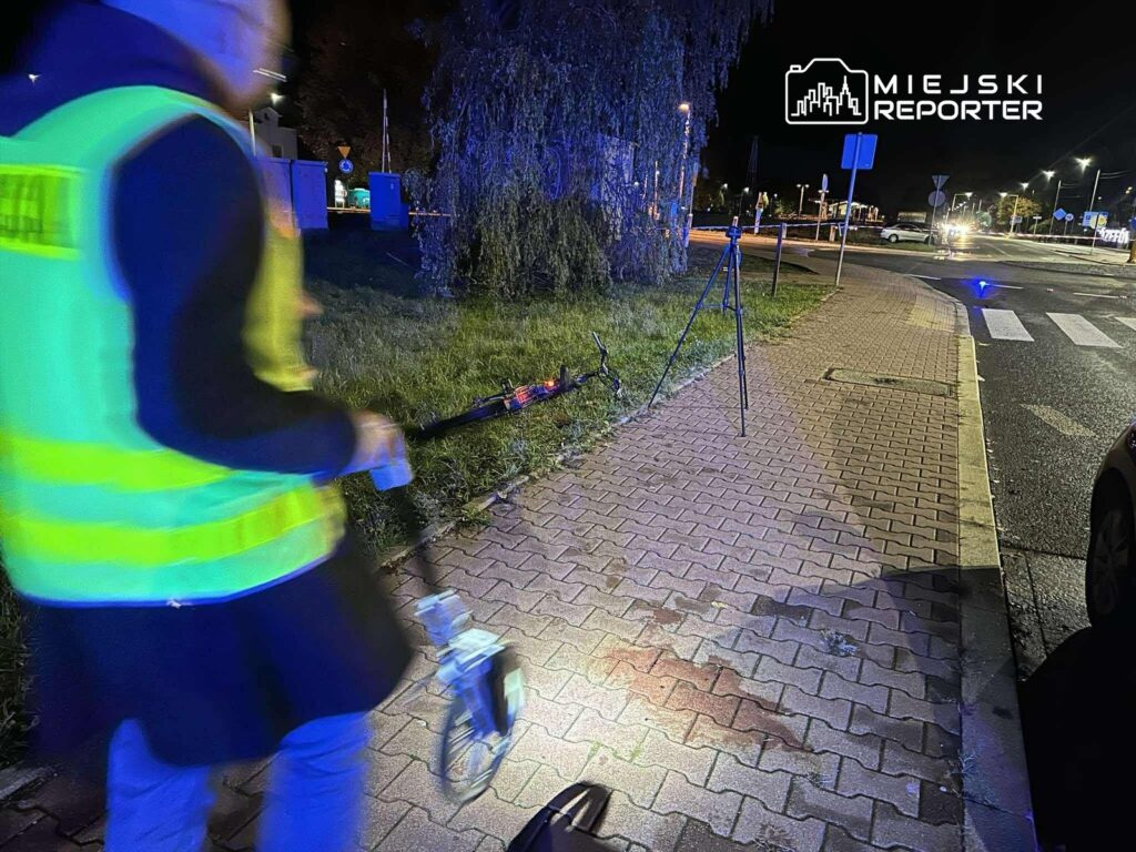 Mężczyzna w odblaskowej kamizelce mierzy odległość przy ulicy nocą, obok statywu i nieoświetlonego terenu.