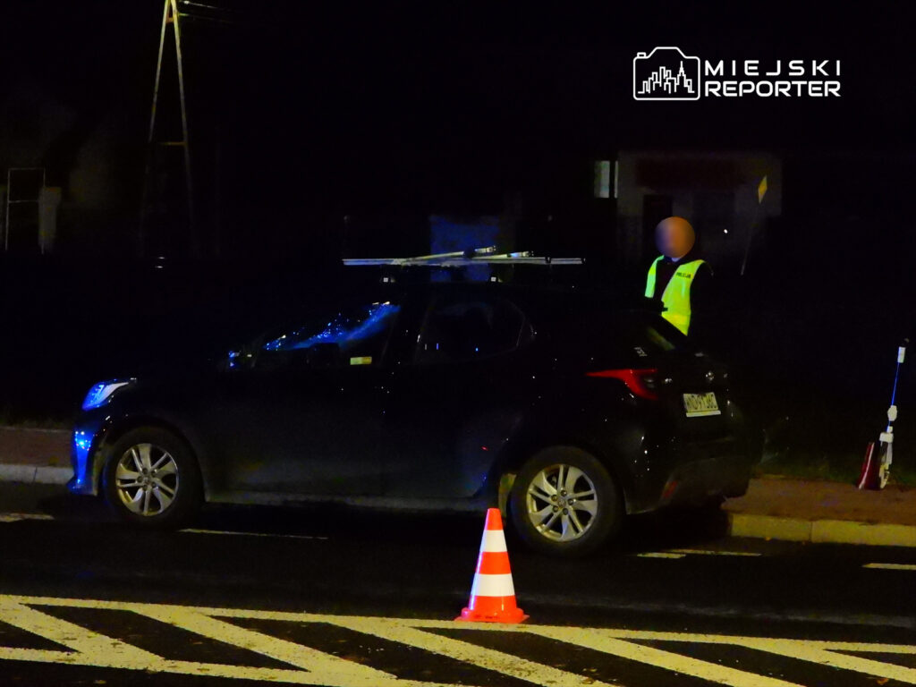 Policjant w odblaskowej kamizelce kontroluje ruch drogowy obok samochodu z włączonymi światłami.