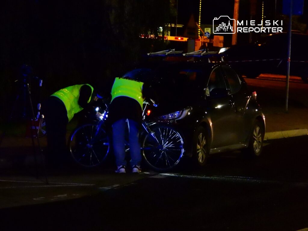 Dwaj policjanci w odblaskowych kamizelkach badają rower obok zaparkowanego samochodu w nocy.