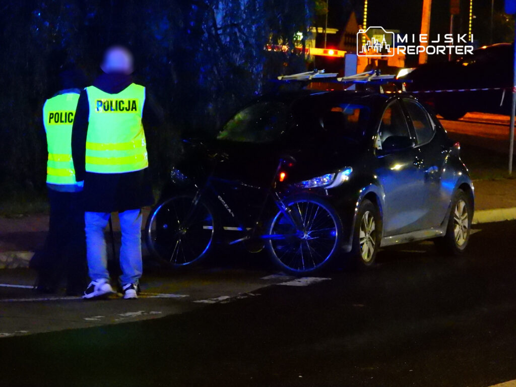 Policjanci w odblaskowych kamizelkach rozmawiają obok czarnego samochodu z rowerem przy drodze w nocy.