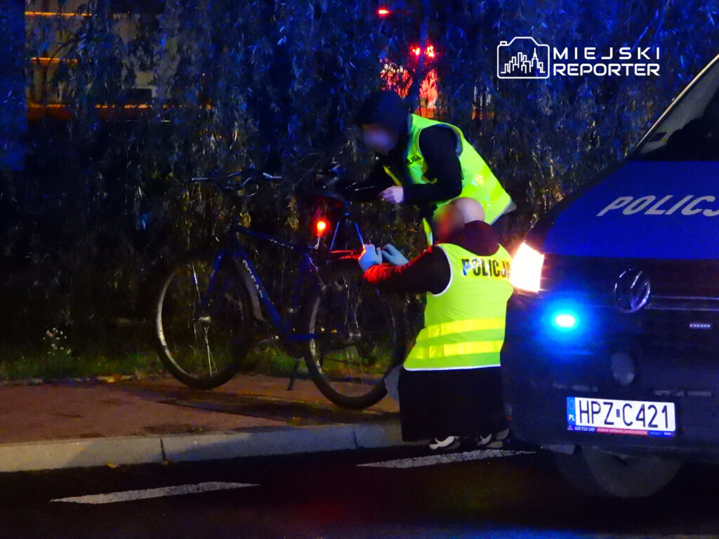 Policjanci w odblaskowych kamizelkach sprawdzają rower na poboczu, obok radiowozu z włączonymi sygnałami świetlnymi.