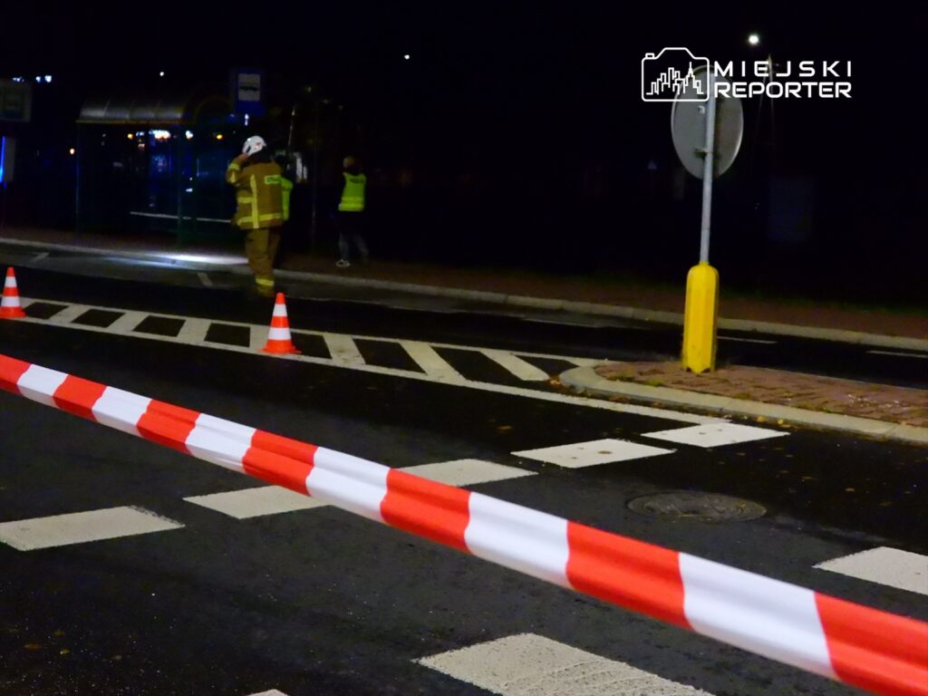 Strażak w mundurze kieruje ruchem na zablokowanej ulicy, oznaczonej taśmą ostrzegawczą.