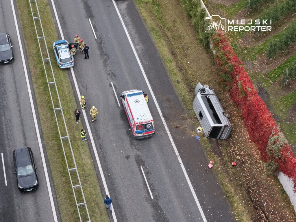 Na drodze leży przewrócony samochód dostawczy, obok stoją strażacy i policja, a w tle widać zjeżdżające pojazdy.