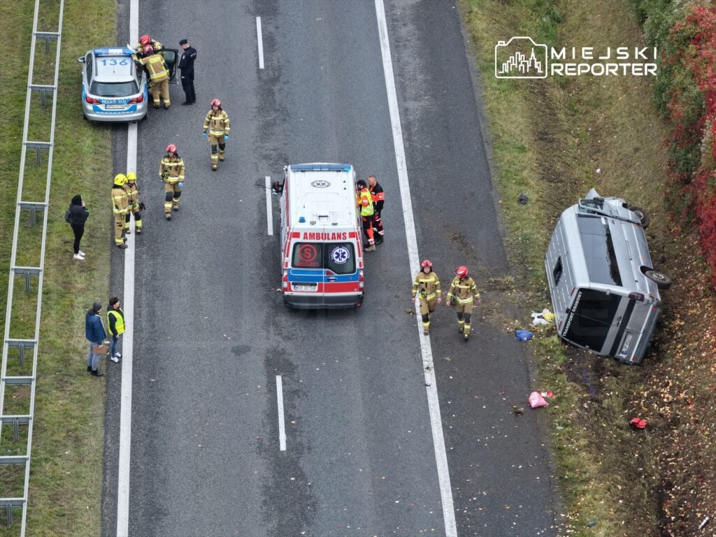 Grupa strażaków i ratowników medycznych pracuje na miejscu wypadku drogowego, obok przewróconego pojazdu.