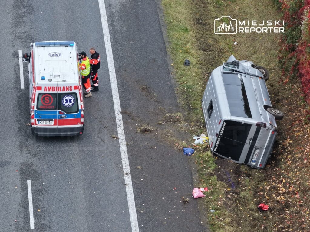 Ratownicy medyczni rozmawiają przy ambulansie obok przewróconego samochodu dostawczego na poboczu drogi.