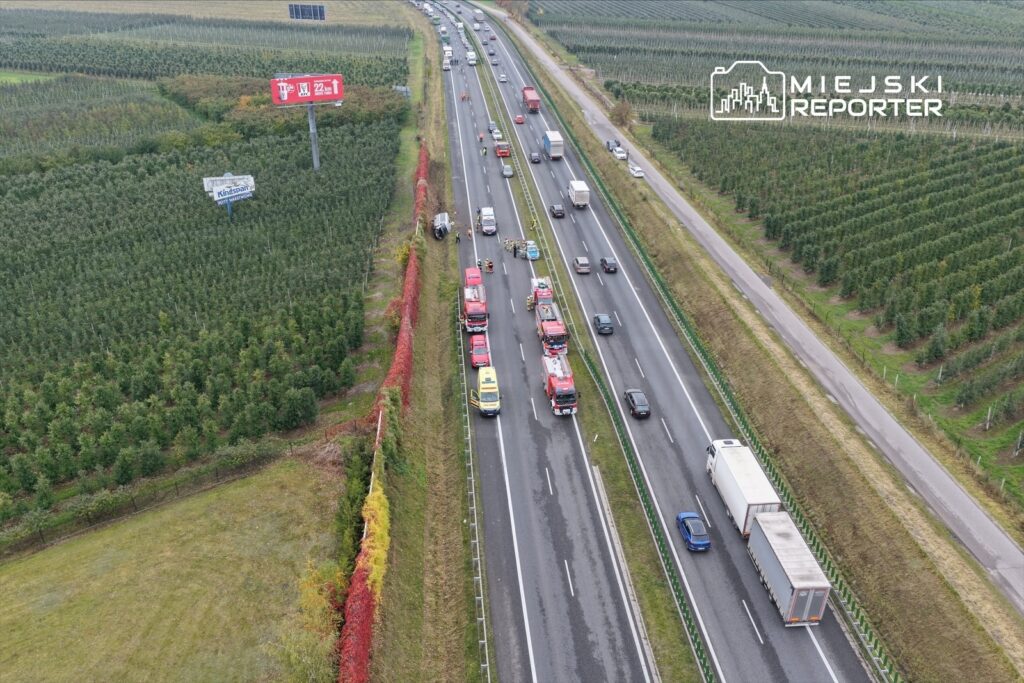 Ruch samochodowy na drodze, z pojazdami ratunkowymi przy barierce, w otoczeniu sadów.